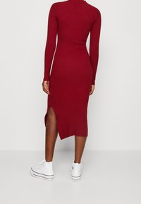 Personne portant une robe midi rouge ajustée à manches longues avec une fente latérale, associée à des baskets blanches montantes Converse, debout devant un fond uni.