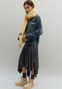 Zöld csíkos hosszú ujjú ing, szürke redős szoknya, sárga sál és bézs bokacsizma műszőrme díszítéssel. Texturált anyag és kényelmes szabás.