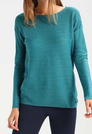 Femme portant un pull côtelé à manches longues bleu sarcelle avec un col rond et un pantalon bleu marine, debout devant un fond blanc.