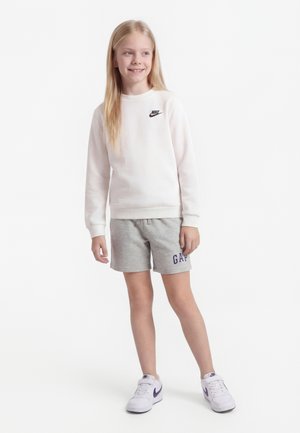 Νεαρή κοπέλα που στέκεται, φοράει λευκό φούτερ Nike, γκρι σορτς GAP και λευκά αθλητικά Nike, χαμογελά και κοιτάζει προς τα πλάγια.