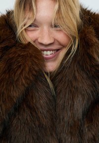 Cappotto in pelliccia sintetica marrone con un aspetto spesso e testurizzato; modella con leggere lentiggini e capelli biondi sorride mentre è avvolta nel capo.