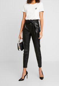 Schwarze Kunstleder-High-Waist-Hosen mit vorderem Bindedetail, kombiniert mit einem weißen Baumwoll-T-Shirt, das eine kleine Schlangengrafik zeigt.