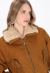 Giacca bomber marrone con colletto beige soffice, chiusura frontale con zip e due tasche laterali con dettagli a bottone. Tessuto dalla superficie liscia.