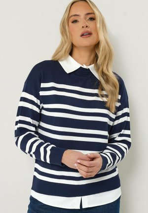 Frau mit langen blonden Haaren, die einen marineblauen und weiß gestreiften Pullover über einem weißen Hemd mit Kragen trägt, steht mit sanft verschränkten Händen.