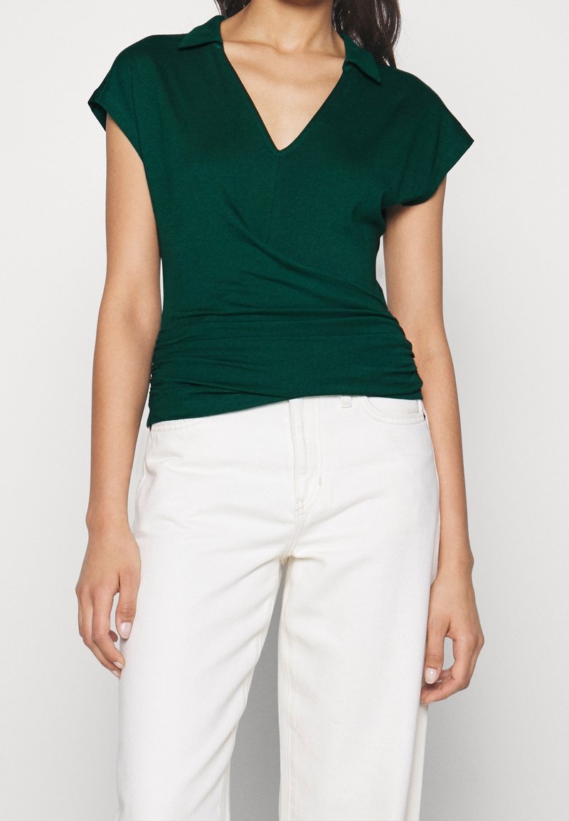 Femme portant un haut vert foncé à manches courtes en V avec col et un pantalon blanc taille haute, debout devant un fond uni.
