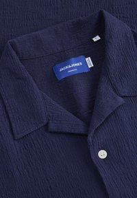 Camisa de color azul marino con cuello y tejido texturizado, que presenta un solo botón blanco y una etiqueta de marca cosida con "JACK & JONES" visible.