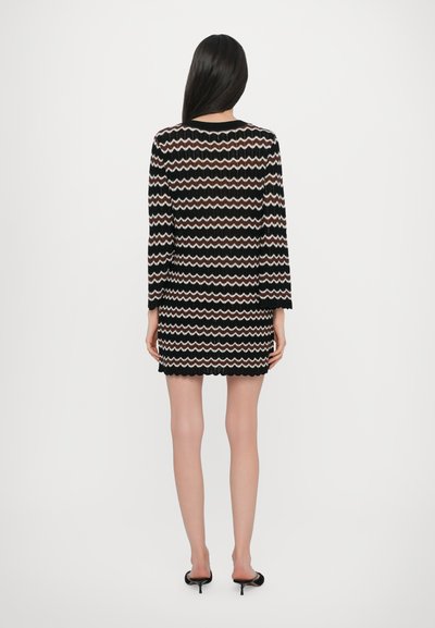 Robe courte noire et crème avec un motif zigzag, texture tricotée, manches longues et ourlet à volants. Associée à des escarpins noirs à bout ouvert.