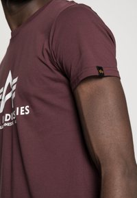 Maroon short-sleeve t-shirt made of smooth fabric, featuring white logo and design detail on the front, with a black tag accent on the sleeve.