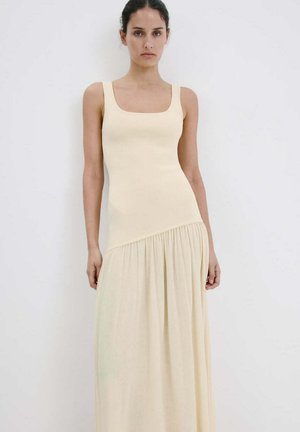 Mujer con vestido maxi crema sin mangas con escote cuadrado y falda fruncida, de pie sobre fondo blanco liso.