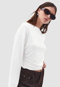 Bílý top s dlouhým rukávem, bočními řasením, kulatým výstřihem a měkkou bavlněnou tkaninou, kombinovaný s hnědými kalhotami se metalickými akcenty.