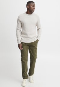 Hellbeiger strukturierter Pullover, grüne gerade geschnittene Hose und weiße Sneakers. Das Model steht mit den Händen in den Taschen und schaut leicht zur Seite.