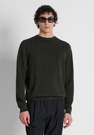 Giovane uomo con capelli ricci che indossa un maglione nero a maniche lunghe con motivo, pantaloni neri e occhiali da sole rettangolari scuri, in piedi davanti a uno sfondo semplice.