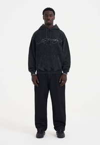 Black hooded sweatshirt with front pocket, featuring white embroidered brand name. Paired with loose-fitting black pants and black sneakers.