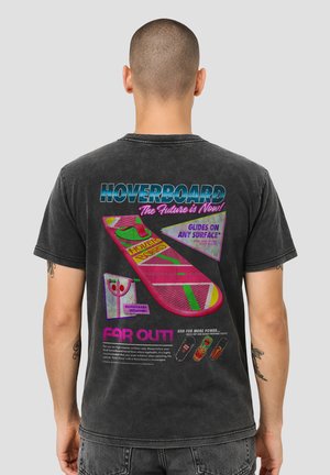 Man wearing black t-shirt with colorful retro hoverboard design and text on the back promoting a futuristic hoverboard product.