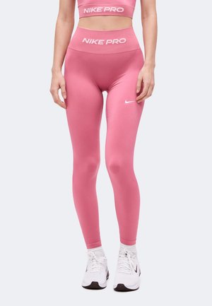Femme portant un legging Nike Pro rose et un soutien-gorge de sport assorti avec des baskets blanches Nike, debout devant un fond uni.