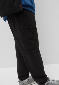 Schwarze Cargo-Hosen mit mehreren Taschen, einem lockeren Schnitt und Reißverschluss-Details, kombiniert mit grauen Sportschuhen.