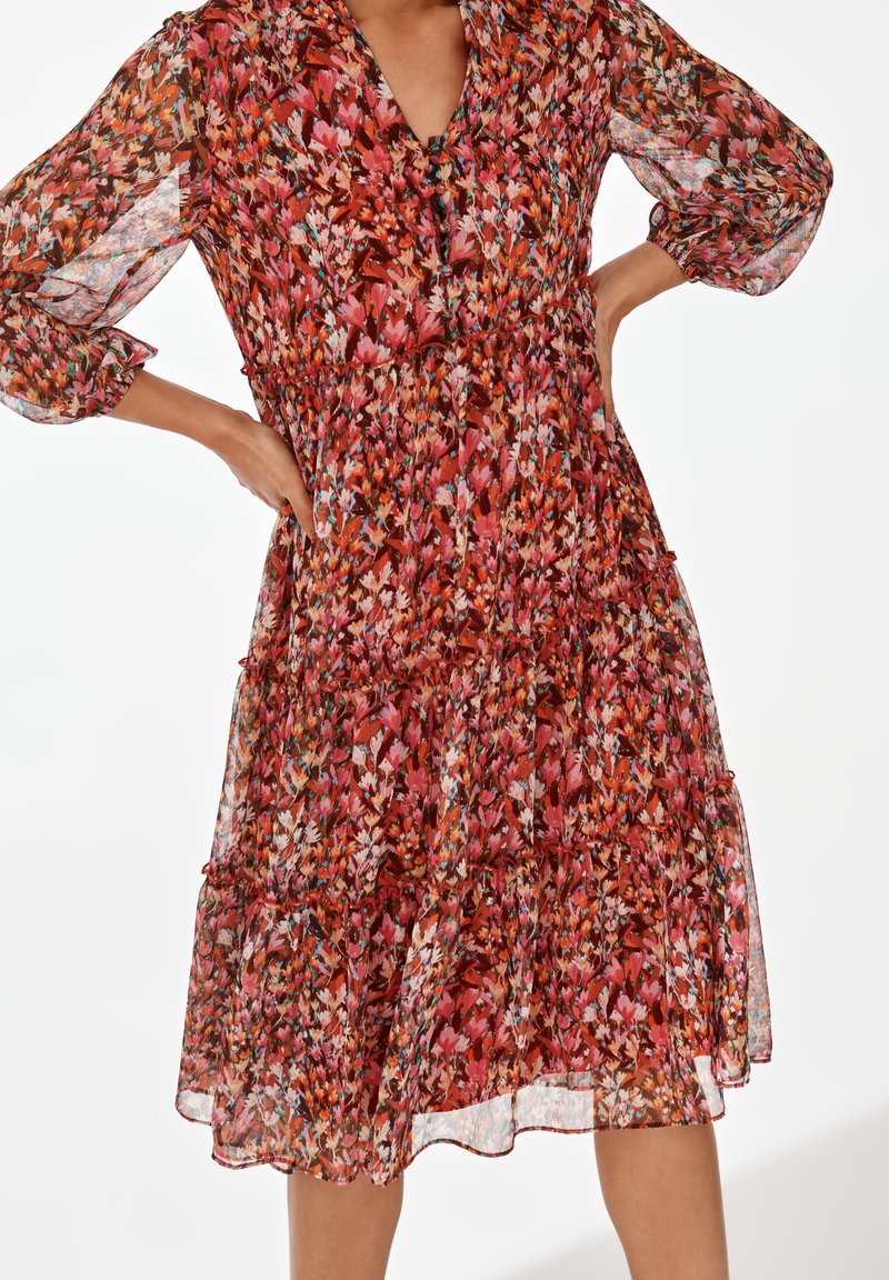 Femme portant une robe longue jusqu'aux genoux à manches longues avec un motif floral rouge, rose et orange et un col en V.