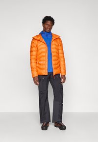 Casaco puffer laranja com capuz, apresentando uma textura leve. Camada intermédia azul com fecho éclair e calças pretas com bolsos. Sapatos pretos texturizados.