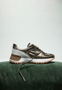 Zapatilla metálica bronce con acentos blancos, parte superior texturizada, diseño de cordones y suela de goma. Presentada sobre un fondo texturizado verde.