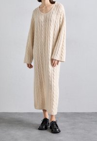 Person klädd i en lång krämfärgad kabelstickad klänning med långa ärmar och svarta snörskor, stående på ett grått golv.