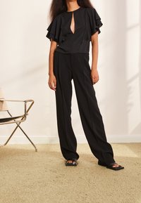 Top nero con maniche a farfalla e dettaglio a chiave abbinato a pantaloni neri a gamba larga. Il modello indossa sandali neri flat. Tappeto beige texture.