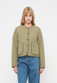 Oliwkowa pikowana kurtka z okrągłym dekoltem, zapięciami na napy, dwoma przednimi kieszeniami i czarnymi wykończeniami. Stylizowana z niebieskimi dżinsami.