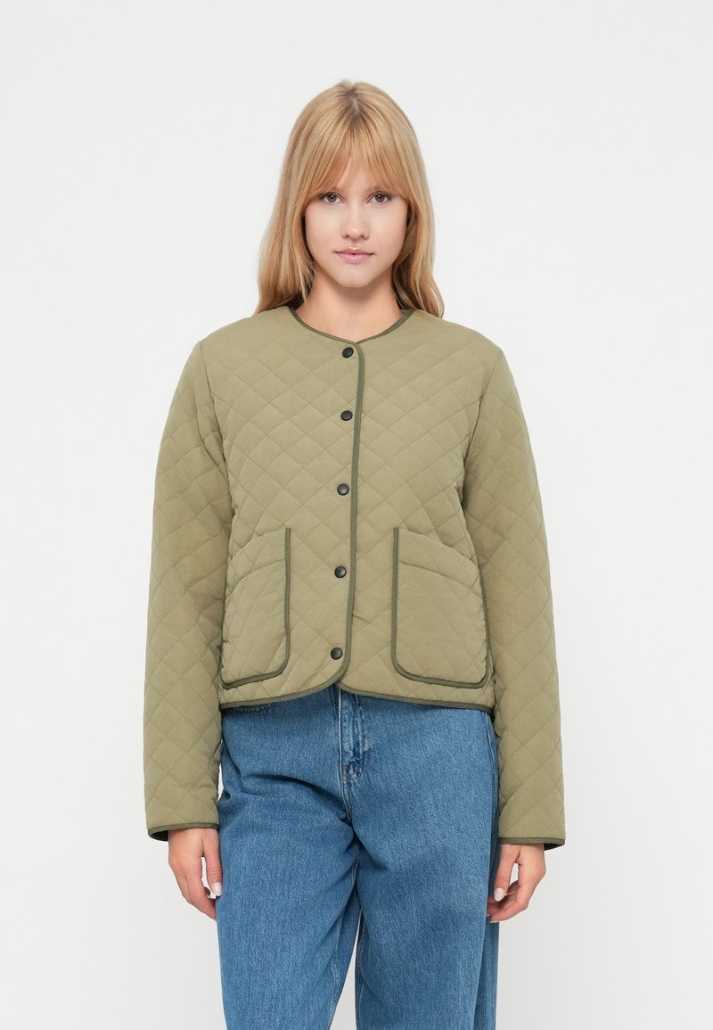 Oliwkowa pikowana kurtka z okrągłym dekoltem, zapięciami na napy, dwoma przednimi kieszeniami i czarnymi wykończeniami. Stylizowana z niebieskimi dżinsami.
