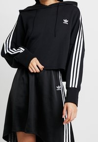 Svart croppad hoodie med vitt Adidas-logotyp och tre vita ränder på ärmarna, tillsammans med en svart satin kjol med en liten Adidas-logotyp.