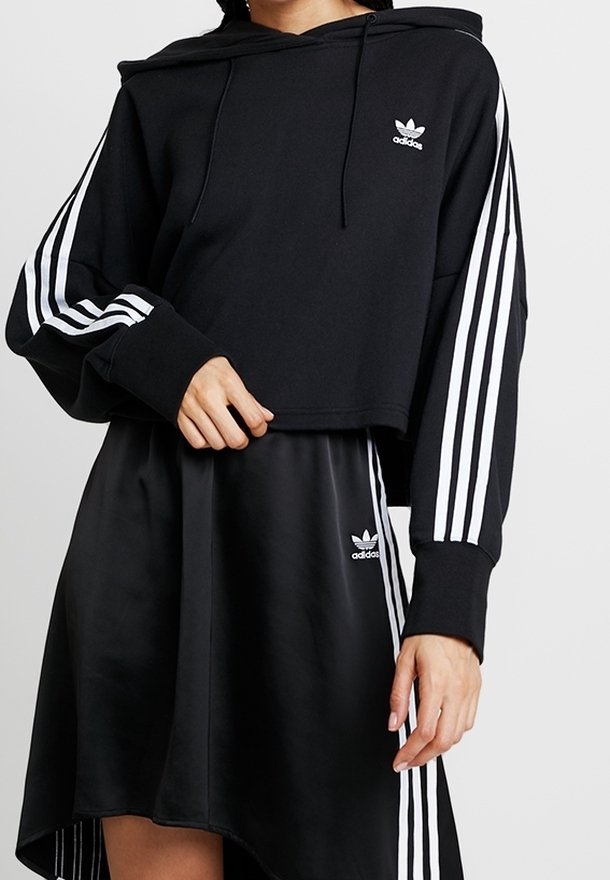 Svart croppad hoodie med vitt Adidas-logotyp och tre vita ränder på ärmarna, tillsammans med en svart satin kjol med en liten Adidas-logotyp.