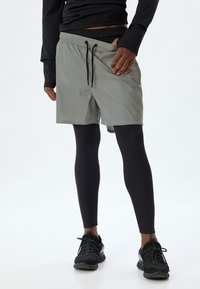 Lys grå træningsshorts med elastisk talje, sort snor og sideslids, parret med sorte kompressionsleggings og sneakers.