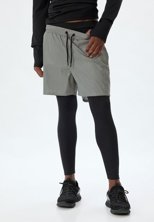 Lys grå træningsshorts med elastisk talje, sort snor og sideslids, parret med sorte kompressionsleggings og sneakers.