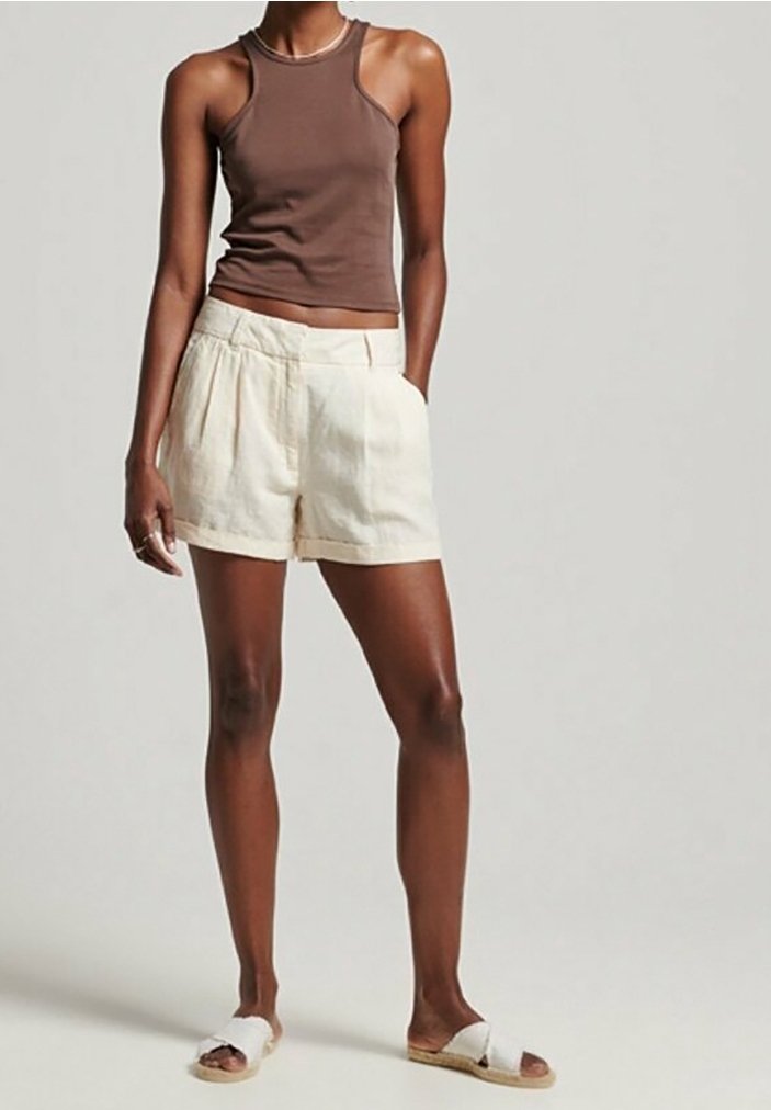 Femme portant un débardeur marron sans manches, un short beige taille haute avec des poches et des sandales blanches à enfiler, debout devant un fond uni.