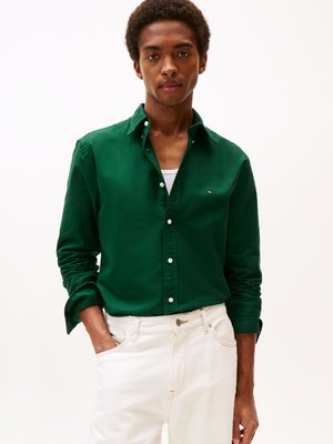 Camicia verde a maniche lunghe con tessuto testurizzato e colletto, abbinata a jeans bianchi. La camicia presenta un piccolo logo e le maniche arrotolate.