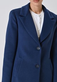 Donna che indossa un cappotto blu navy su misura con ampie revers e due bottoni sopra una camicia bianca con bottoni rivestiti in tessuto.
