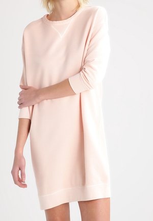 Mujer con un vestido sudadera de manga larga de color rosa claro, de pie frente a un fondo blanco con un brazo cruzado sobre el otro.