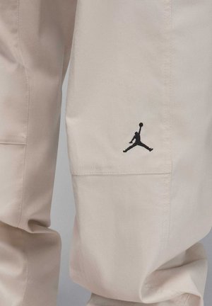 Beige cargo bukser lavet af letvægtsstof, med et sort broderet Jumpman-logo tæt på lommen og synlige sy detaljer.