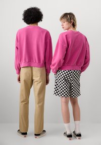 Två personer står rygg mot rygg, klädda i rosa sweatshirts, en med beige byxor och den andra i svart-vit rutiga shorts.