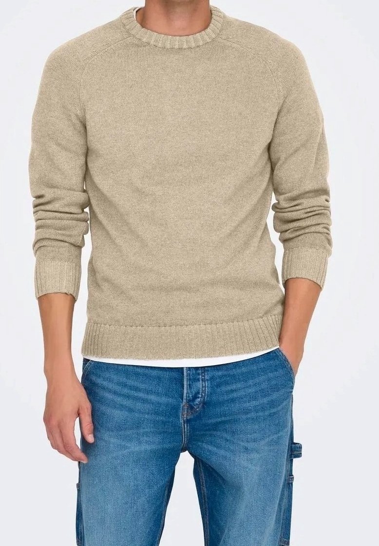 Homme portant un pull beige en maille avec des poignets et un ourlet côtelés, un jean bleu et un sous-vêtement blanc, debout avec une main dans la poche.