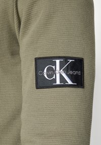 Olíva zöld, bordázott anyagú, hosszú ujjú felső, fekete folttal, amelyen fehér betűkkel a "CK" és a "Calvin Klein Jeans" felirat látható az ujjon.