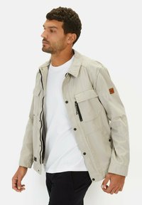 Lys beige jakke lavet af stof, med en frontlynlås, flere lommer og et patch på venstre ærme. Designet med en krave.