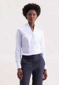 Camicia azzurra a righe con colletto alla cinese, maniche lunghe e un piccolo dettaglio del logo. Abbinata a pantaloni scuri.