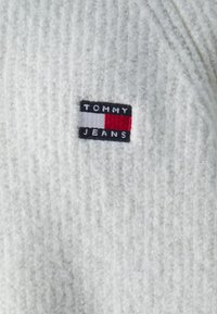 Fehér bordázott pulóver texturált felülettel, fekete, piros és fehér színű téglalap alakú "TOMMY JEANS" logóval.
