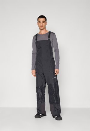 Zwarte waterdichte overalls met verstelbare schouderbanden, ritszakken en textuurpanelen op de benen, gecombineerd met een grijze top met lange mouwen en zwarte schoenen.