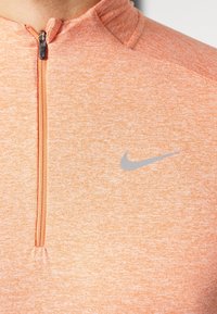 Lähikuva oranssista neulemaisesta urheilupaidasta, jossa on lyhyt vetoketju ja harmaa Nike-logon swoosh rintakehässä.
