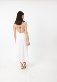 Abito bianco lungo fino ai piedi con design scollato sulla schiena e sottili spalline. Presenta un piccolo dettaglio decorativo sul retro, abbinato a sandali alti leggeri.
