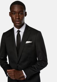 Černý oblek na míru s výpustkovým límcem, bílá košile a černá kravata. Obsahuje kapesníček, hladkou látku a přiléhavý design.