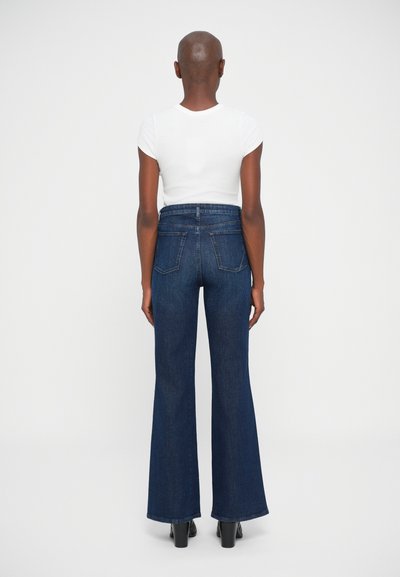 Persona con cabeza rapada, vestida con una blusa blanca de manga corta, jeans oscuros de tiro alto y acampanados, y zapatos de tacón negros, de pie de espaldas.