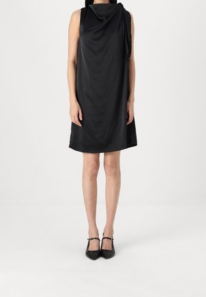 Kobieta w czarnej, satynowej sukience bez rękawów, sięgającej do kolan, z marszczonym wysokim kołnierzem i czarnych, szpiczastych butach typu Mary Jane na obcasie.