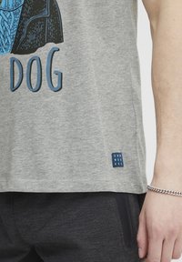 Grå bomulls t-skjorte med blå grafisk design og teksten "DOG." Inkluderer en liten blå etikett med teksten "DNW WORKRS" på siden.