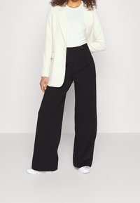 Svarta vida byxor med en slät textur, ihop med en ljus krämfärgad kavaj och en vit skjorta, fullbordat med vita sneakers.